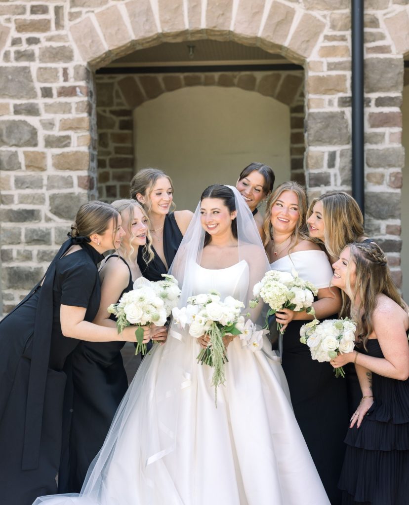 Bride surrounded by bridesmaids in mismatched black dresses holding white bouquets at stone archway