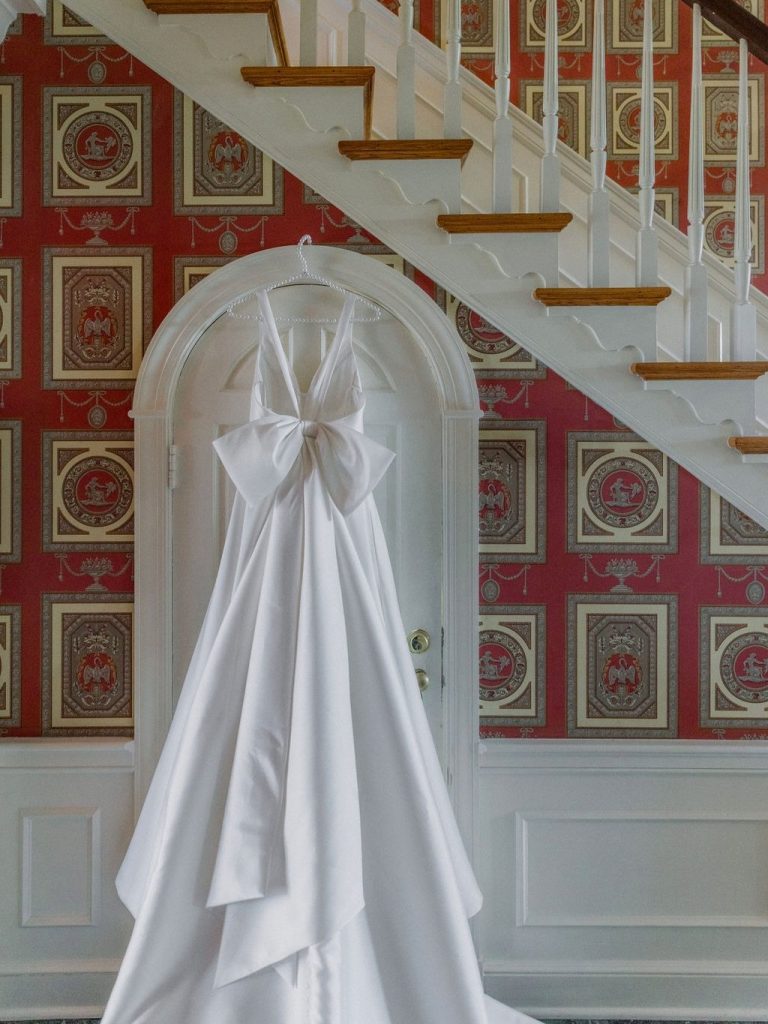 White wedding gown with dramatic bow detail hanging on door beneath staircase with red patterned wallpaper
