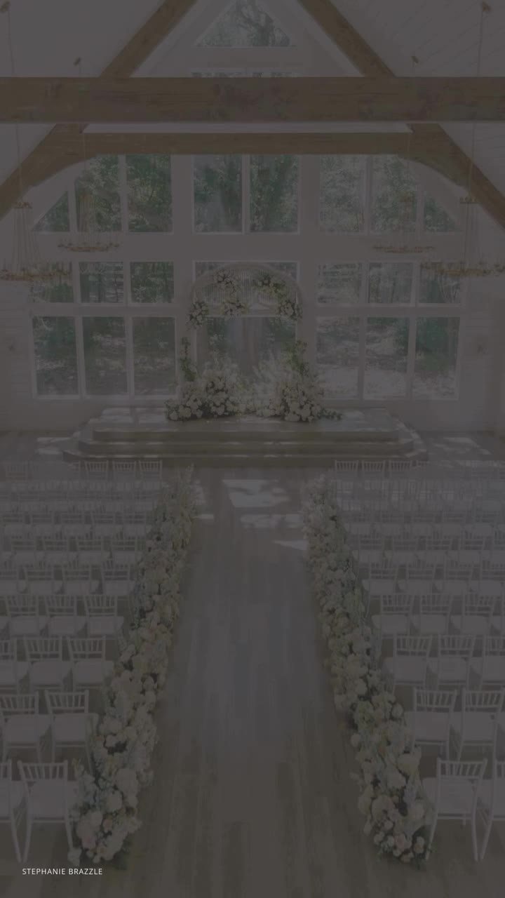 Elegant barn wedding ceremony venue with white floral arch, floor-to-ceiling windows, and rows of white chairs