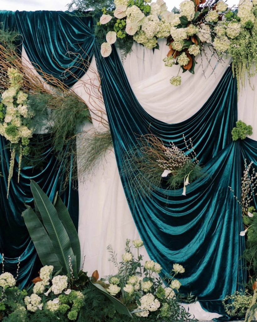 Elegant wedding ceremony backdrop with draped teal velvet and ivory fabric, adorned with white hydrangeas, greenery, and tropical leaves