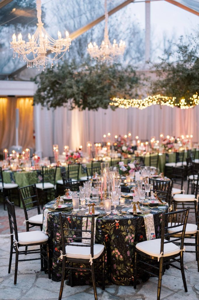 Elegant tented wedding reception with crystal chandeliers, black floral tablecloths, and candlelight at dusk