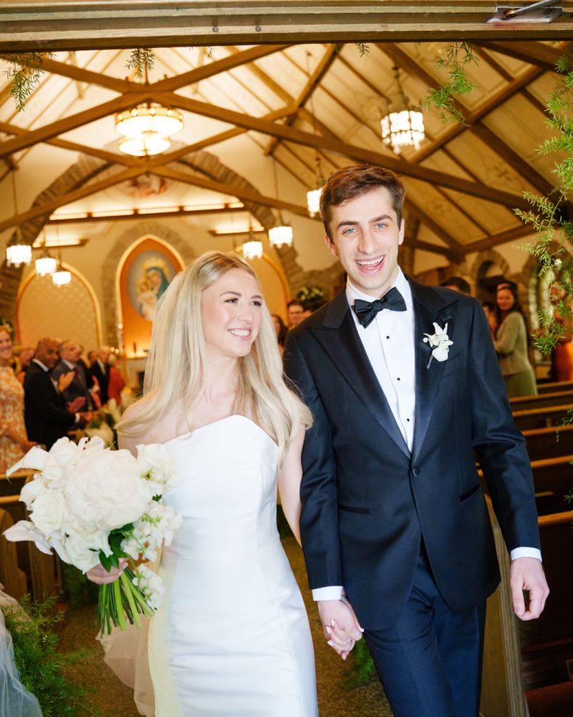 Newlyweds walk down church aisle hand in hand, smiling, with vaulted wooden ceiling and guests in background
