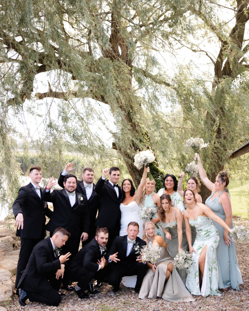 Hannah and Cole's wedding party posing playfully under willow trees with groomsmen in black tuxedos and bridesmaids in sage green dresses