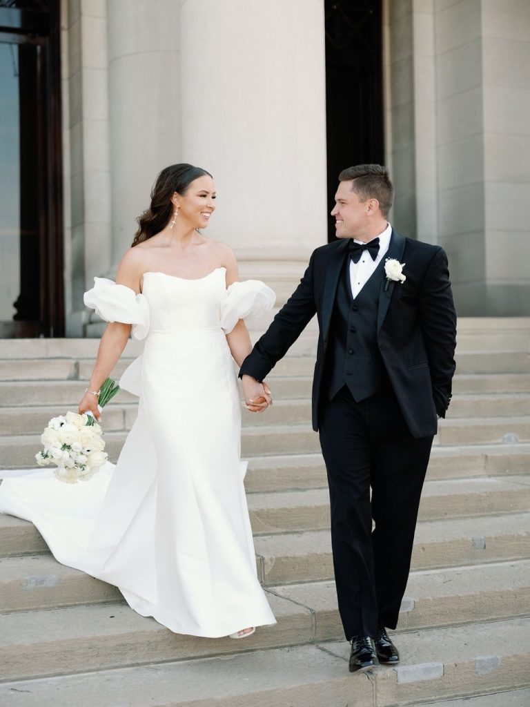 McKenzi and Alex walking hand-in-hand on stone steps, bride in off-shoulder gown, groom in black tuxedo