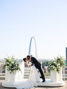 Bride and groom kissing on outdoor ceremony platform with Gateway Arch and white floral arrangements in background