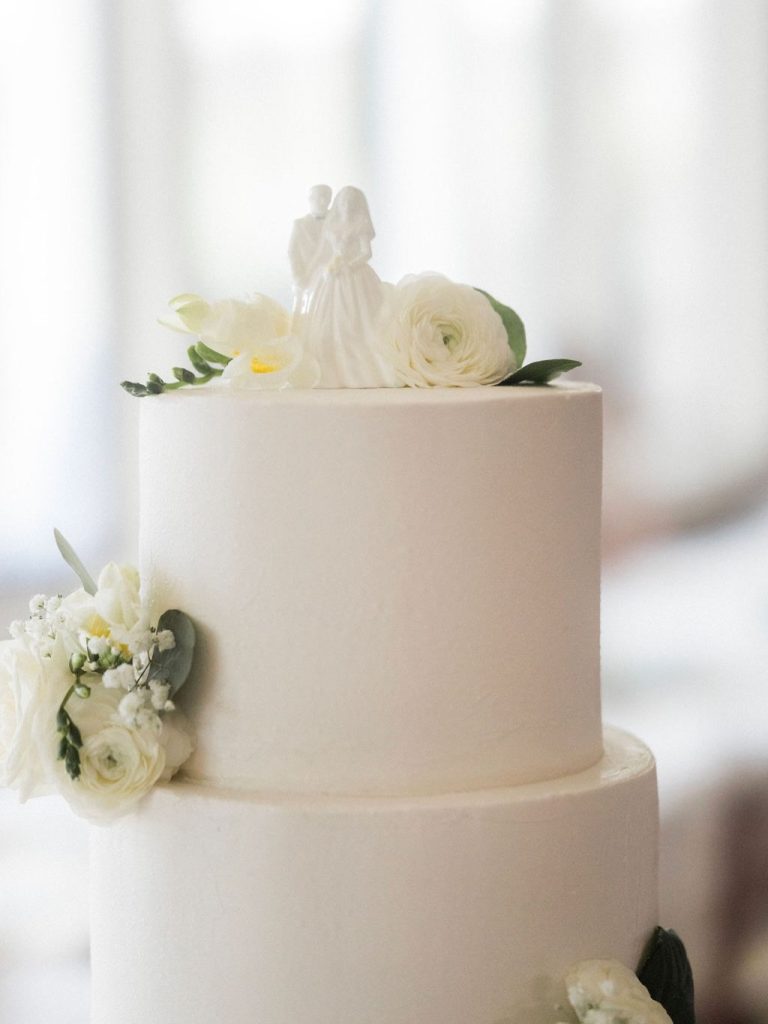 Simple white two-tier wedding cake decorated with white ranunculus and greenery, topped with bride and groom figurine