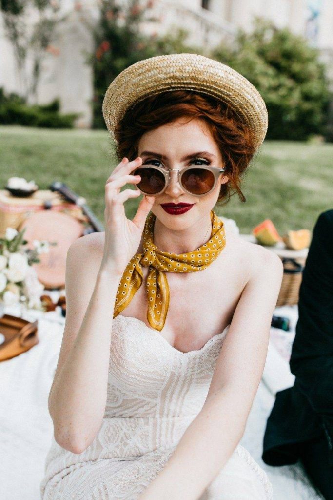 Bride in straw boater hat, yellow polka dot neck scarf, sunglasses, and red lipstick at outdoor wedding