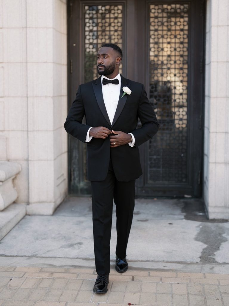 Anthony in black tuxedo with white boutonniere standing in front of ornate doors