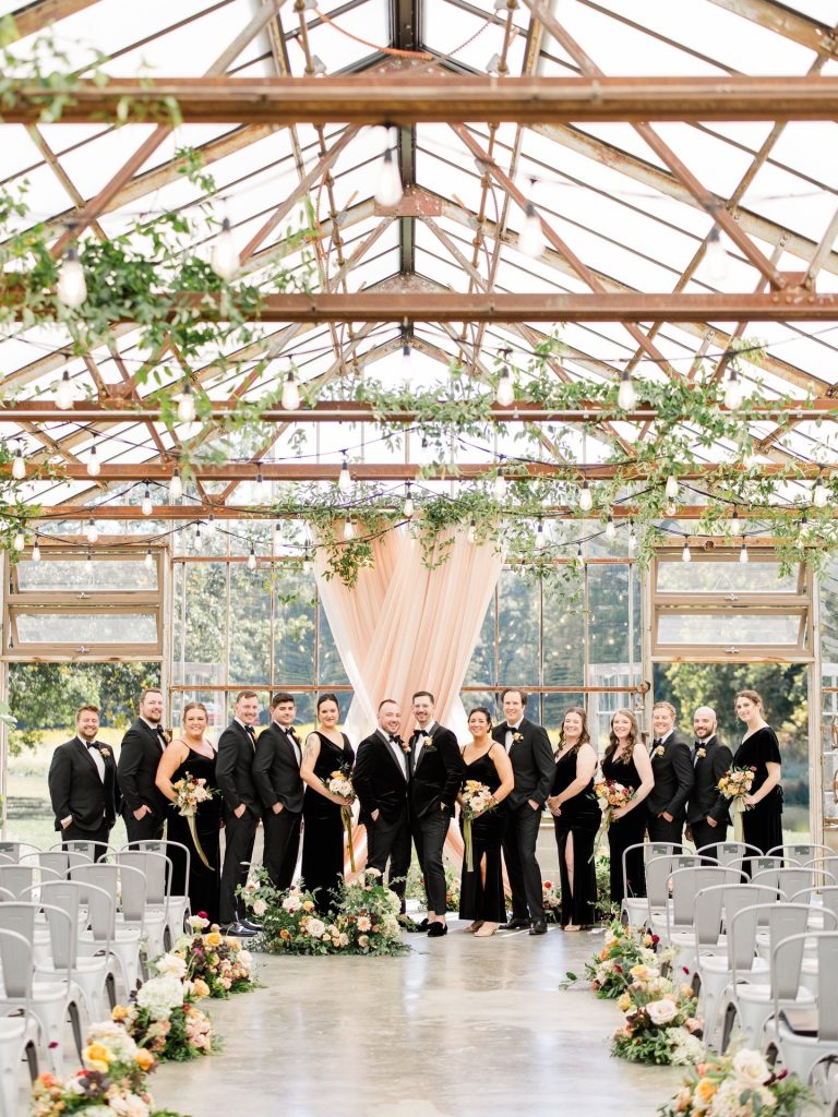 Wedding party in black attire poses beneath exposed wooden beam ceiling and flowing peach drapes in bright glass greenhouse ceremony space at Jorgensen Farms