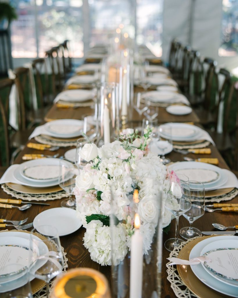 Long wooden reception table set with white plates, gold chargers, tall white candles, and lush white and blush floral centerpieces