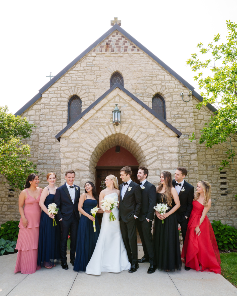Marielle, Kyle, and wedding party in formal attire stand outside stone church with arched entrance