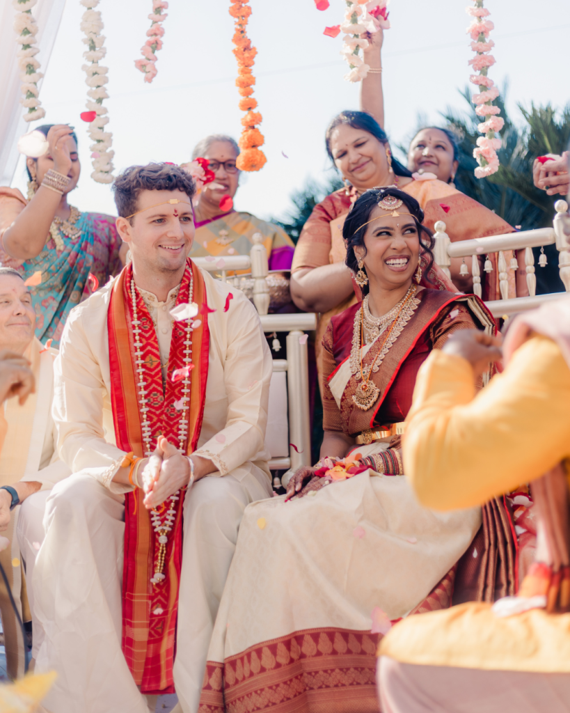 Joyful couple during outdoor Indian wedding ceremony surrounded by family and colorful floral decorations