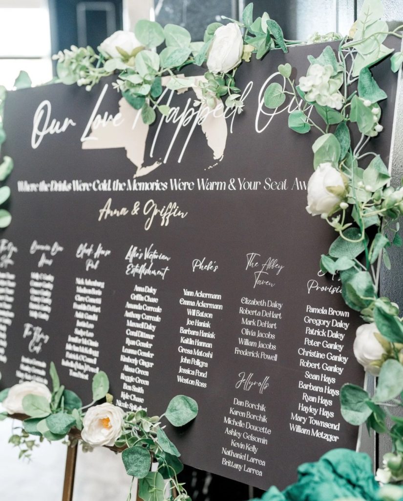 Black acrylic seating chart decorated with eucalyptus and white tulips reading "Our Love Happiest Day"