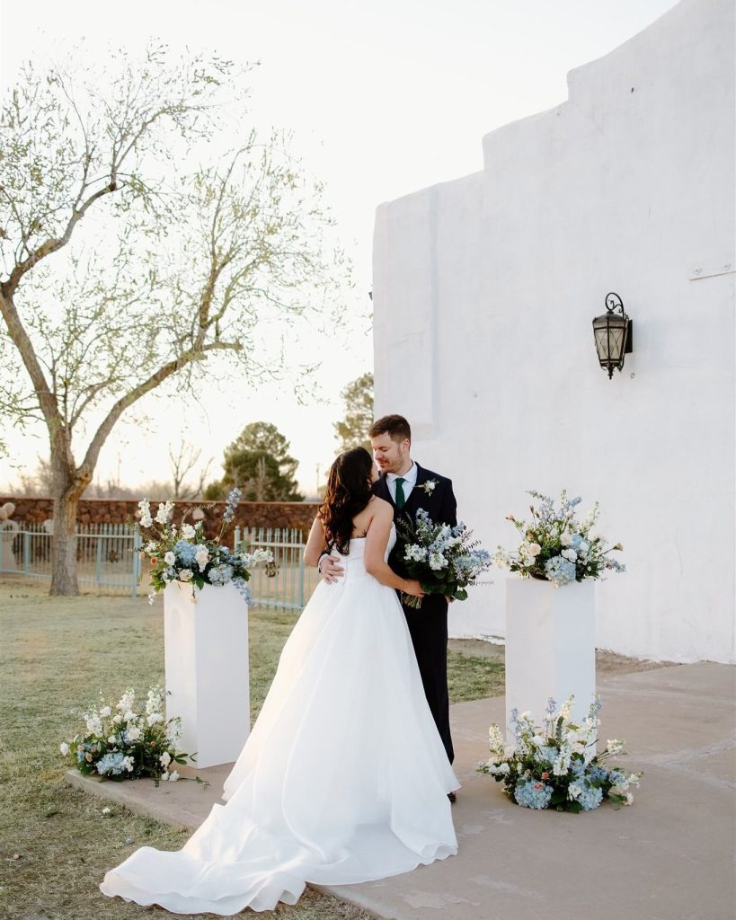 Bride and groom kissing outdoors at white stucco venue with blue and white floral arrangements on pedestals