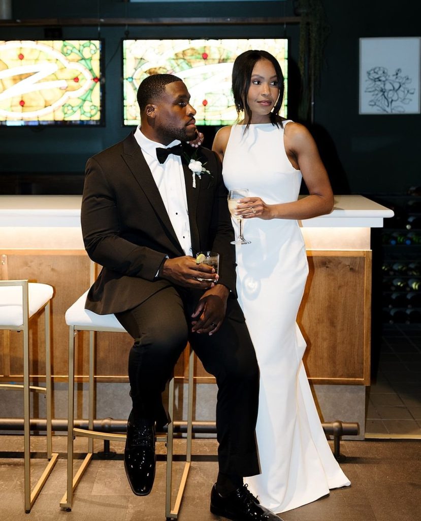 Bride and groom pose with cocktails at modern bar with illuminated stained glass backdrop at The Elliot