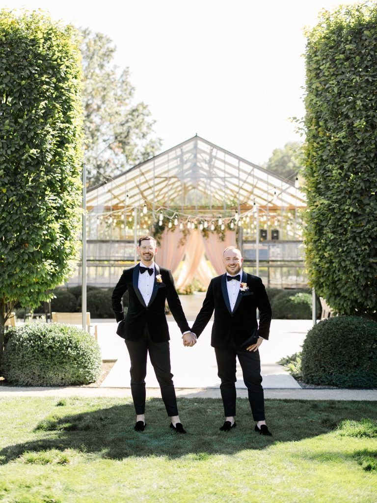 Stephen and Austin hold hands on the grounds of Jorgensen Farms with the glass greenhouse ceremony venue behind them