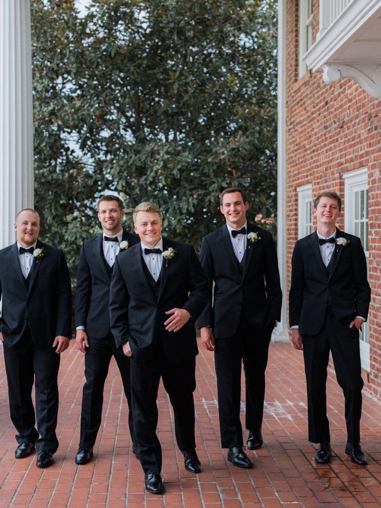 Groom Zach and groomsmen in black tuxedos with bow ties and white boutonnieres standing on brick patio