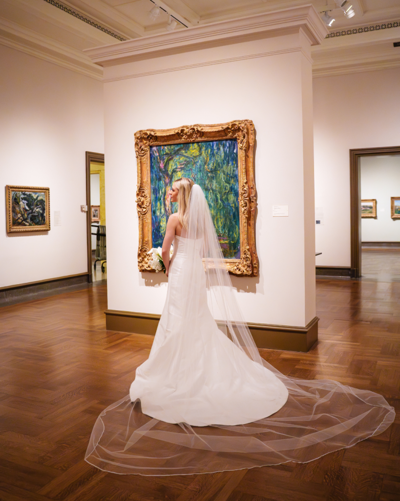 Marielle in wedding gown and cathedral veil stands before ornate framed artwork in museum gallery