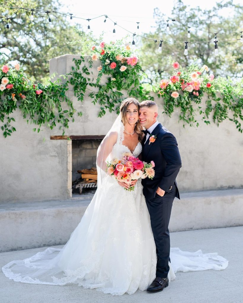 Bride and groom embracing outdoors under string lights and greenery with pink floral arrangements