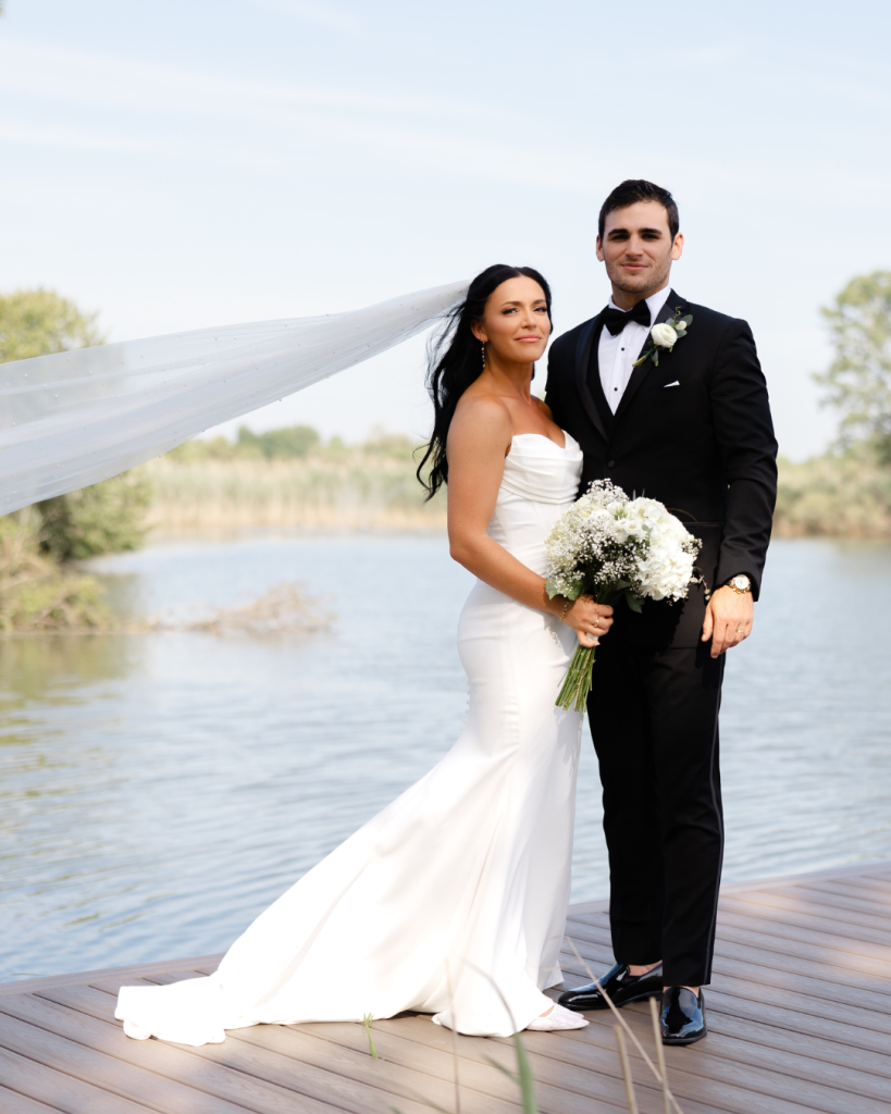 Newlyweds stand together on lakeside dock, bride in strapless gown with white bouquet, groom in classic black tuxedo
