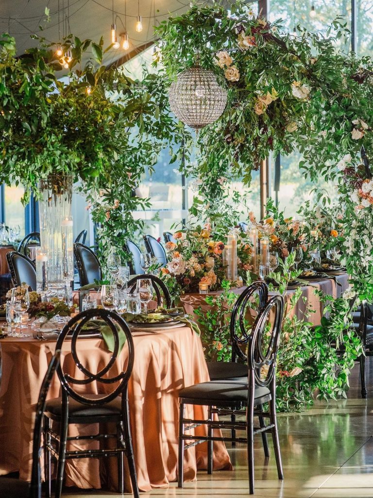 Elegant reception space featuring cascading greenery installations, crystal chandeliers, peach linens, and black Louis chairs at Jorgensen Farms