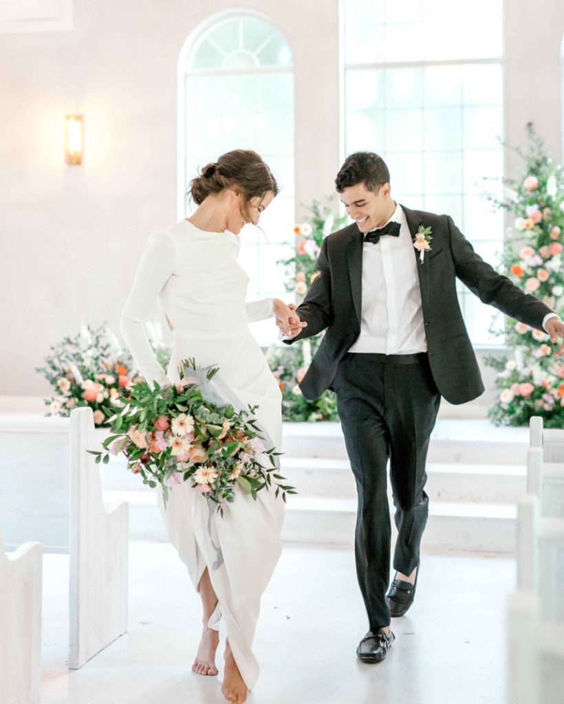 Newlyweds walking hand-in-hand down white chapel aisle holding lush greenery and peach bouquet