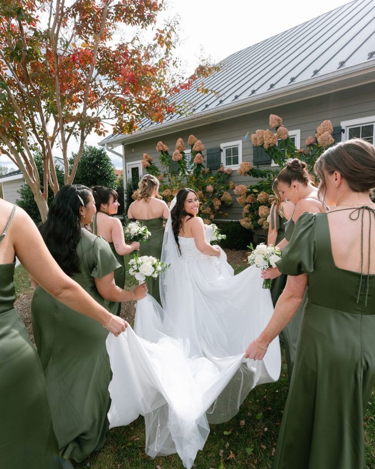 Bridesmaids in sage green dresses help bride with flowing white tulle train in front of rustic barn
