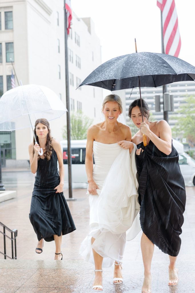 Bride in strapless white gown walks in rain with two bridesmaids in black dresses holding umbrellas on city street
