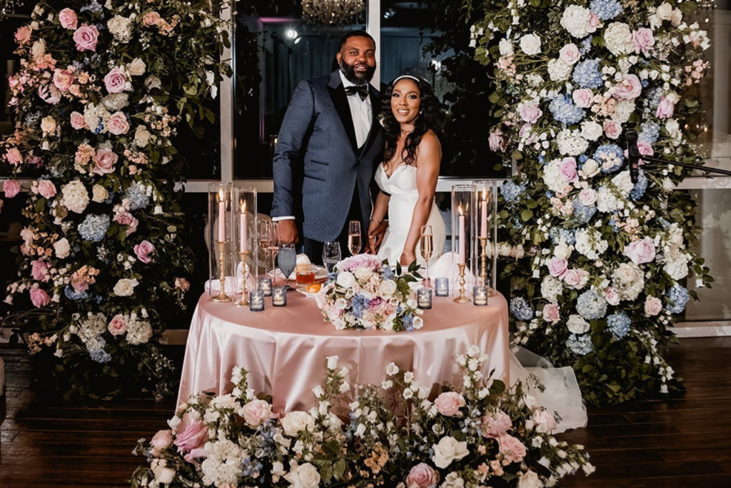 Bride and groom behind sweetheart table with lush pink, white, and blue floral backdrop and romantic candlelit centerpiece