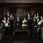 Wedding party in navy formal attire posed in elegant wood-paneled room with white bouquets