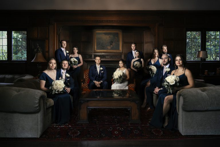 Wedding party in navy formal attire posed in elegant wood-paneled room with white bouquets