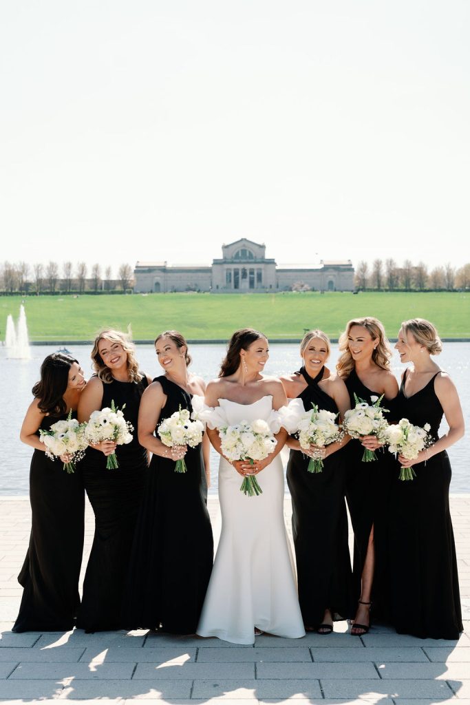 Bride in white gown with six bridesmaids in black dresses holding white bouquets by a reflecting pool
