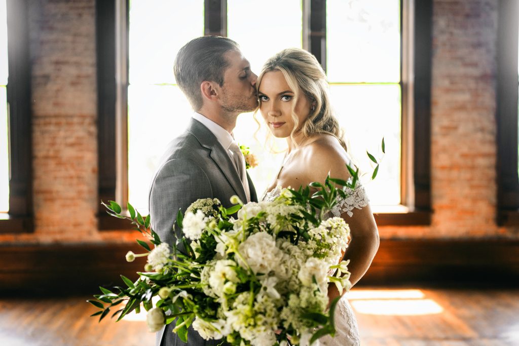 Groom kissing bride's forehead as she holds white and green bridal bouquet in industrial loft venue