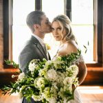 Groom kissing bride's forehead as she holds white and green bridal bouquet in industrial loft venue