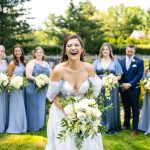 Laughing bride holding white floral bouquet with bridesmaids and groomsmen in blue attire in garden