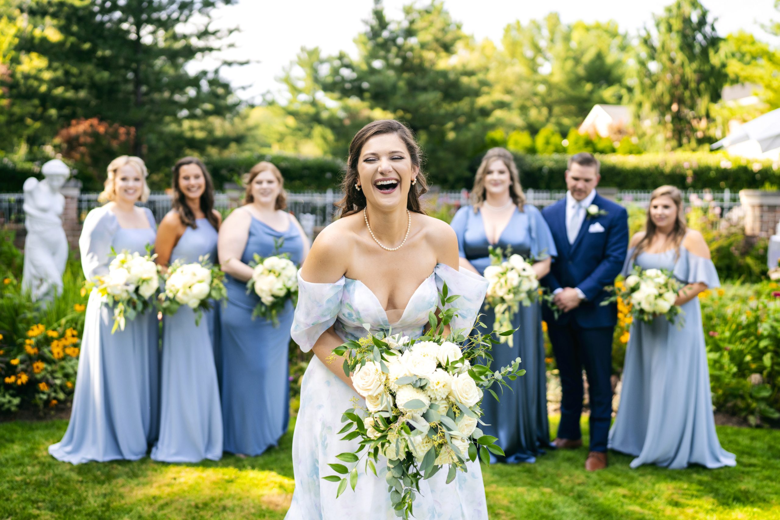 Laughing bride holding white floral bouquet with bridesmaids and groomsmen in blue attire in garden
