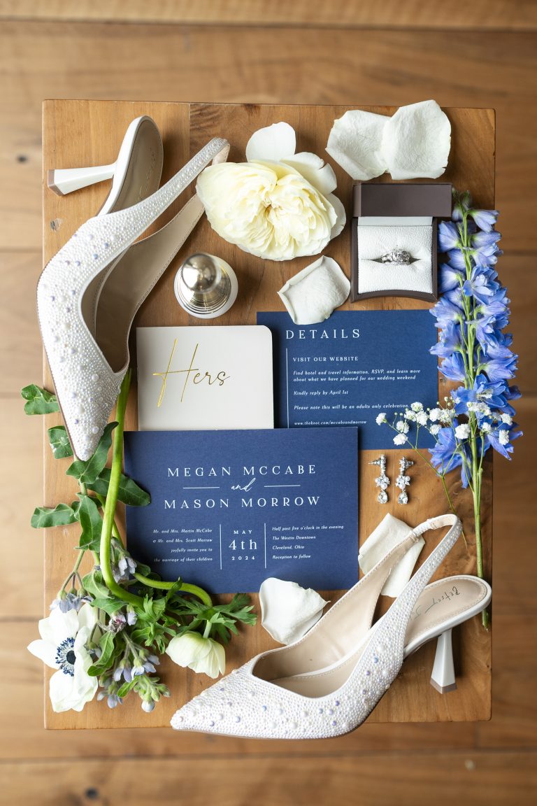Flat lay of wedding details including navy invitation, embellished heels, engagement ring, and blue and white flowers