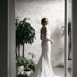Bride in flowing satin gown with train poses in industrial loft with white brick walls and greenery
