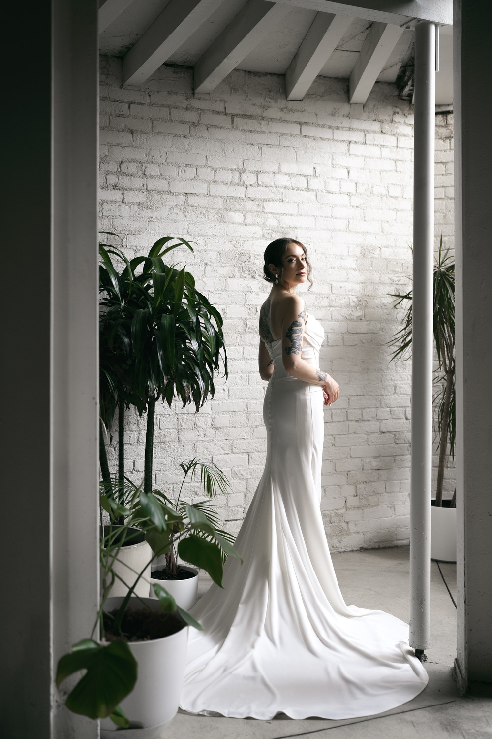 Bride in flowing satin gown with train poses in industrial loft with white brick walls and greenery