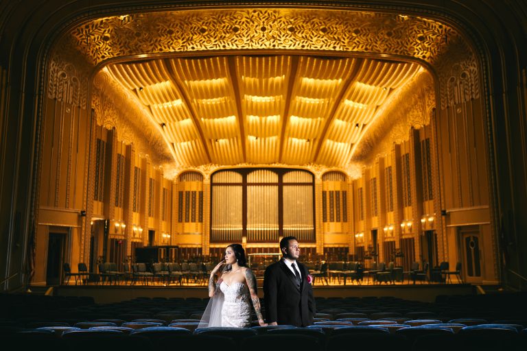 Bride and groom on stage of ornate Art Deco theater with golden illuminated ceiling