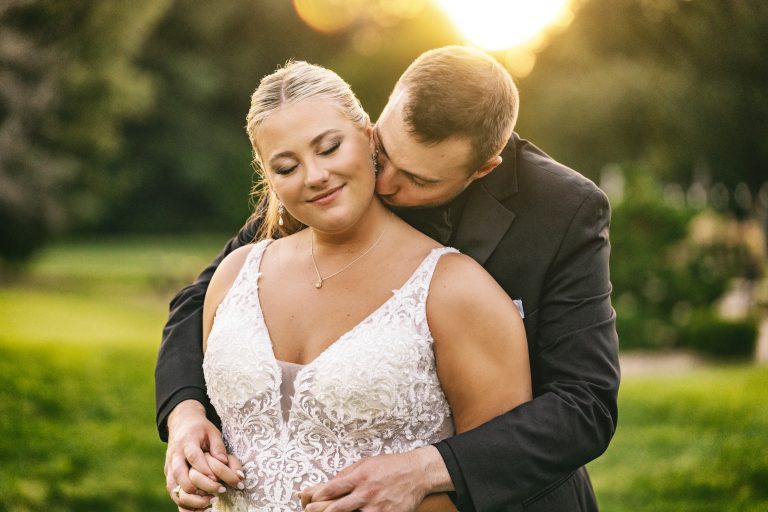 Groom kisses bride's neck as she smiles in lace wedding dress at golden hour