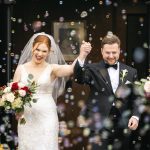 Newlyweds exit ceremony through bubbles, bride holding red and white rose bouquet
