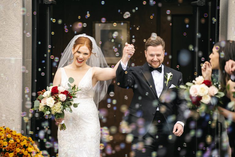 Newlyweds exit ceremony through bubbles, bride holding red and white rose bouquet