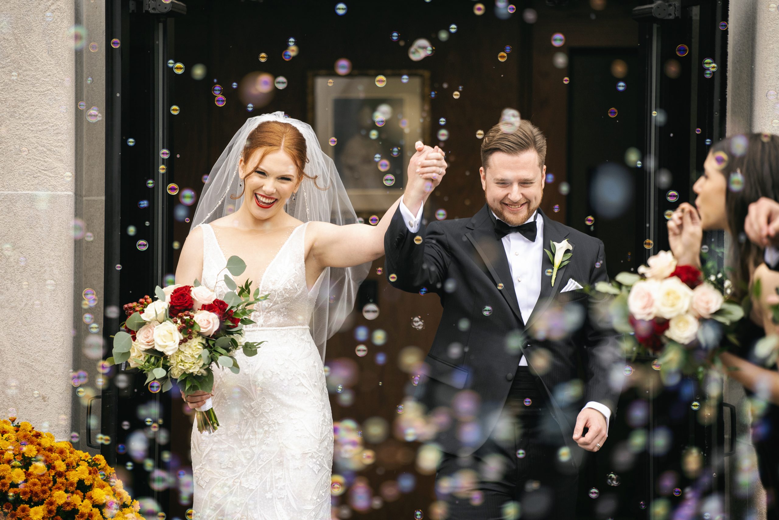 Newlyweds exit ceremony through bubbles, bride holding red and white rose bouquet