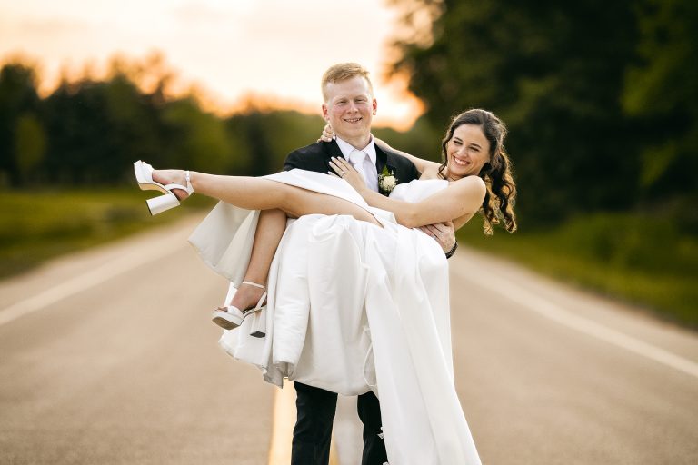 Groom carrying bride on a country road at sunset