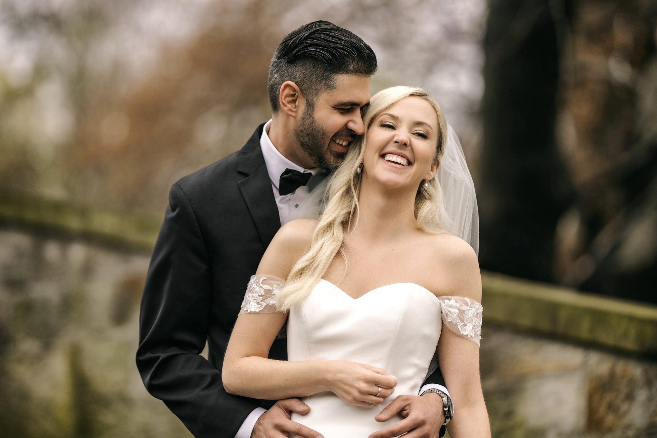 Bride and groom laughing together outdoors, bride in off-shoulder lace wedding gown with veil