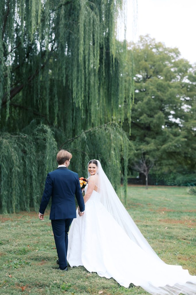 Bride in flowing veil holding colorful bouquet walks hand-in-hand with groom under weeping willow tree