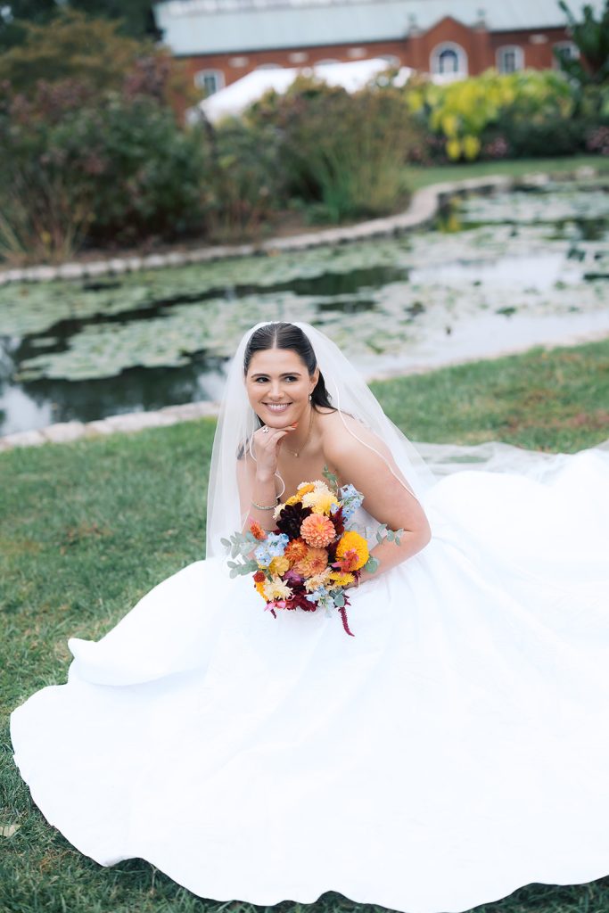 Caroline sits on grass by lily pond in flowing wedding gown holding vibrant multicolored bouquet