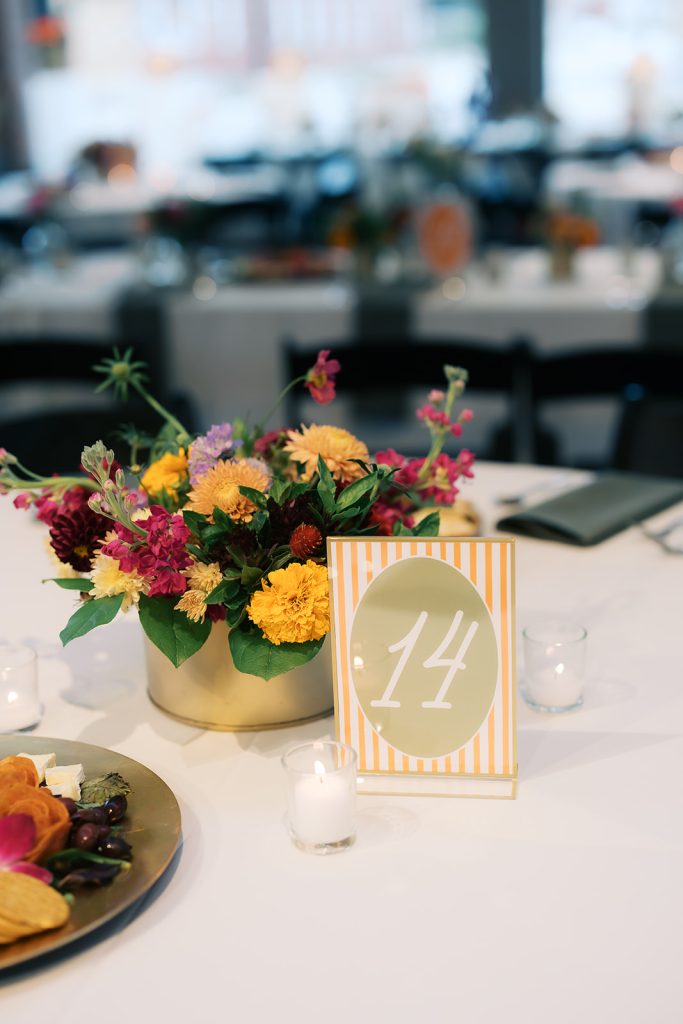 Gold floral centerpiece with orange and pink blooms beside striped table number 14 on white linens