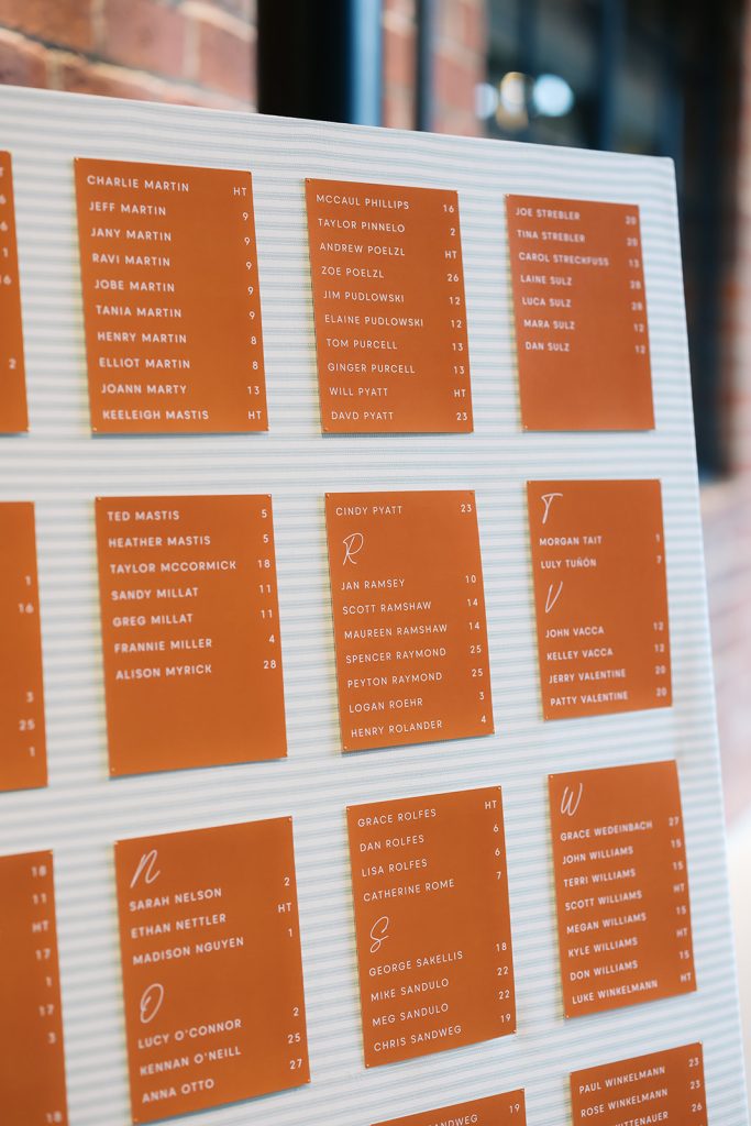 Orange escort cards arranged on white pleated board displaying guest names and table assignments